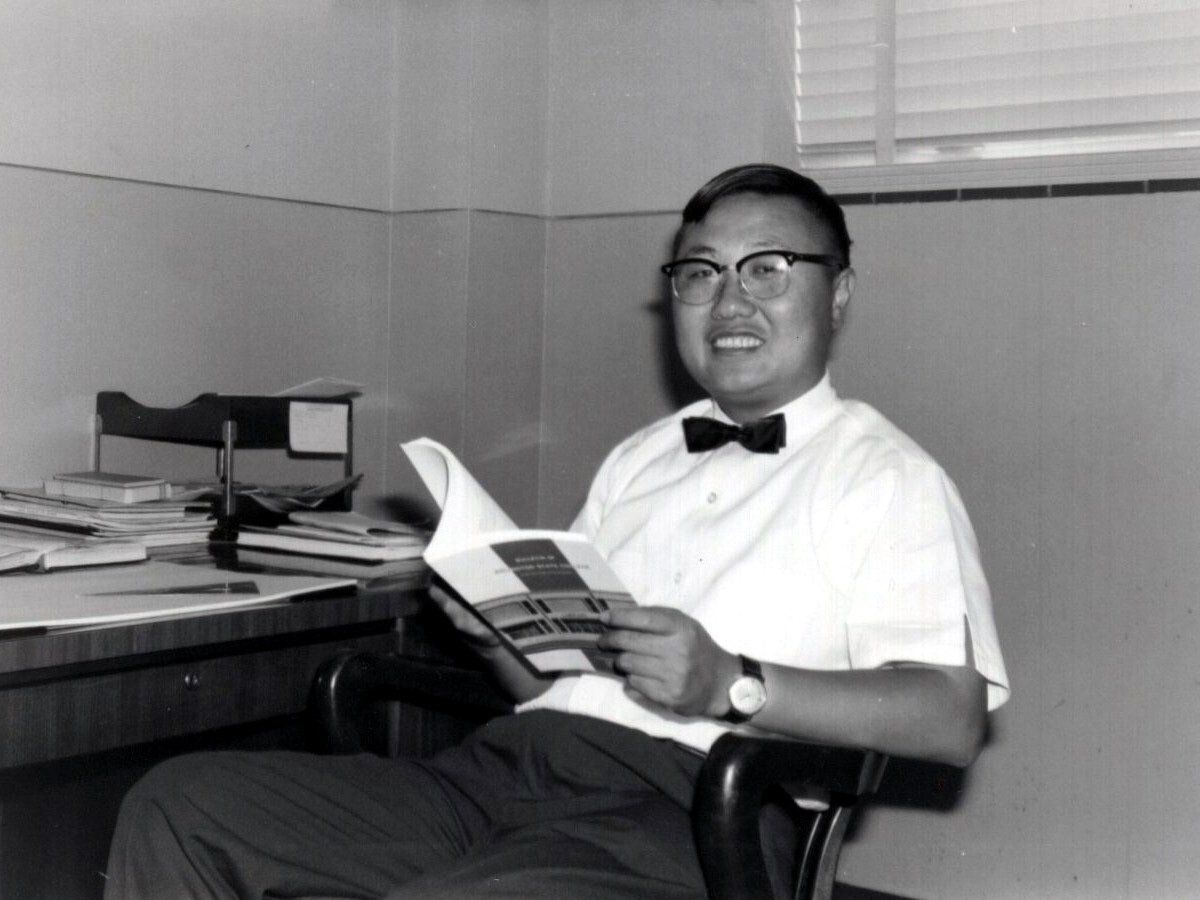 black and white photograph of man wearing a white button-up shirt, bow-tie and glasses while sitting in a chair and smiling at the camera