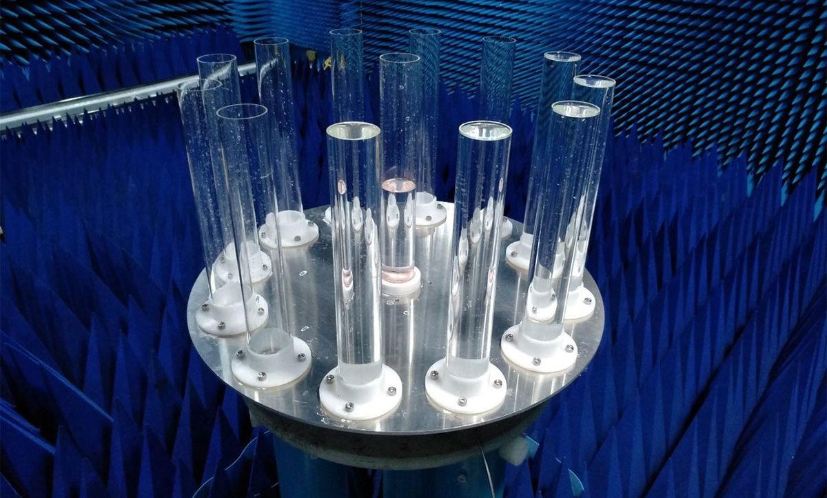 Blue square cones line the walls and floor of a room. On a stand a metal disk lies horizontally. Twelve clear plastic tubes line the circumference, and one tube is in the center. Several of the tubes are filled with water.