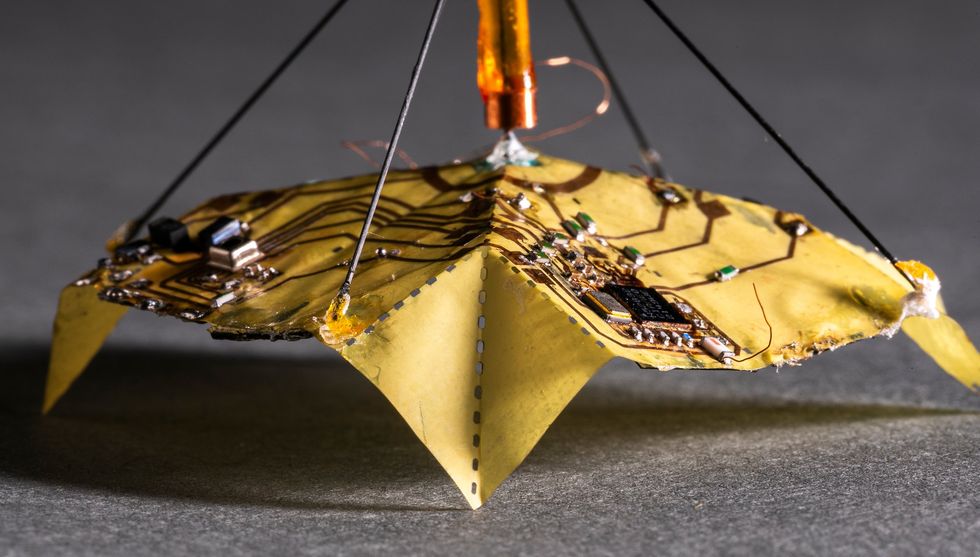 close-up image of a yellow folded origami flier with electronic circuits printed across it and stabilizing wires going from edges of the wing to a central rod that stabilizes the craft