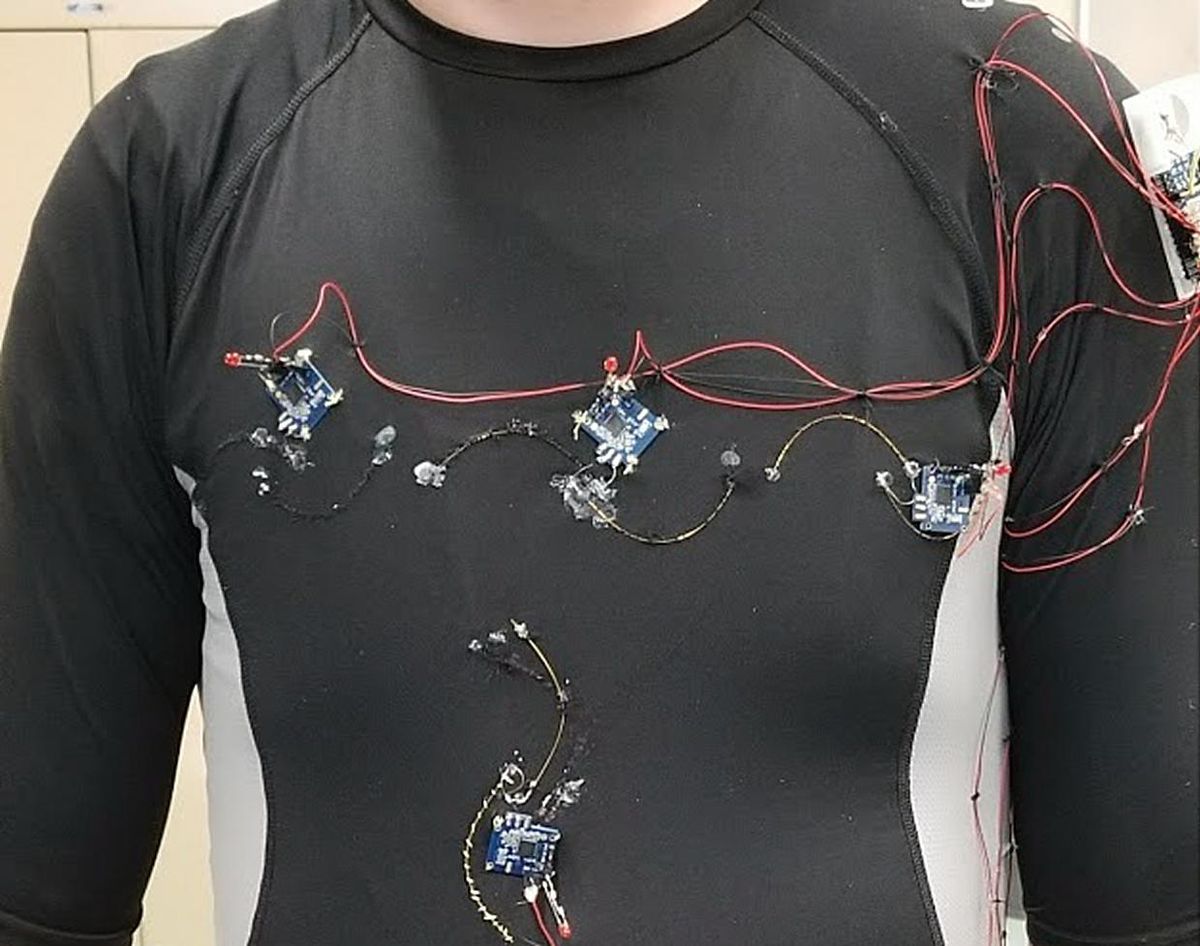 Close up of a person wearing dark long sleeved shirt. Multiple blue sensors and wires cover it.