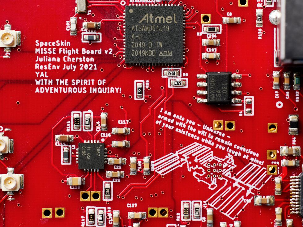 Close up of a red circuit board. Text etched on the board reads u201cSpaceskin MISSE Flight Board v2 Juliana Cherston ResEnv July 2021 YAL With the spirit of adventurous inquiry!u201d and u201cI am onto you u2013 Universe u2013 armed with the will to remain conscious of your existence while you laugh at mine!u201d