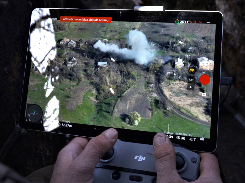 Close up of a tablet screen shows a drone's eye view of a rural scene, with smoke rising from an area. Hands manipulate the DJI drone controller in front of it.