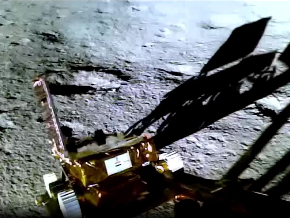 Color TV image of boxy gold rover moving down a ramp away from the camera. The rover's shadow is visible against the gray lunar surface.
