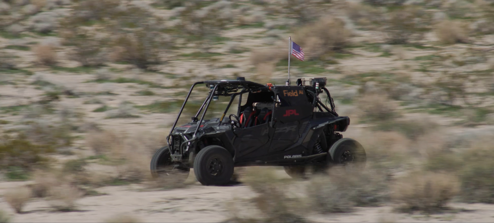 driverless dune buggy-type vehicle with waving American flag drives through a blurred landscape of sand and scrub brush