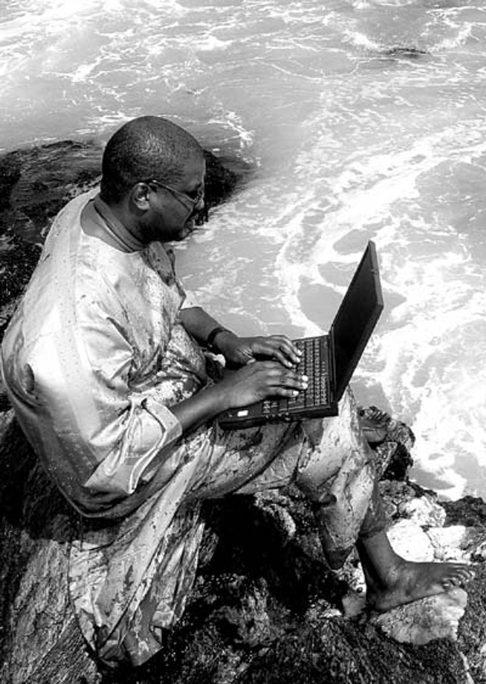 Man holding a laptop, sitting a on a rock near water.  
