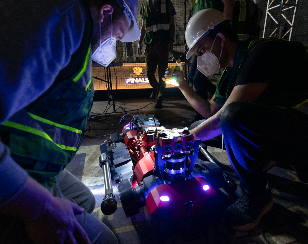 Two roboticists crouch over a red quadrupedal robot in the dark, one holds a flashlight