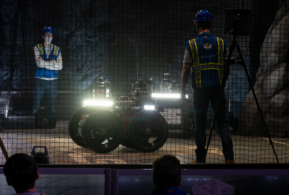 Two roboticists watching three large wheeled robots with bright lights