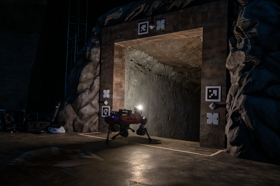 A red quadrupedal robot with a bright light walks into a dark cave entrance