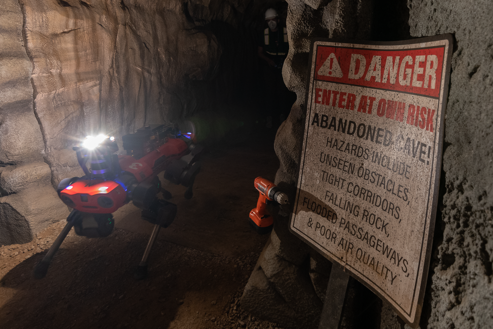 A red quadrupedal robot in a cave walks past a drill on a wall and a sign that says "danger enter at your own risk"