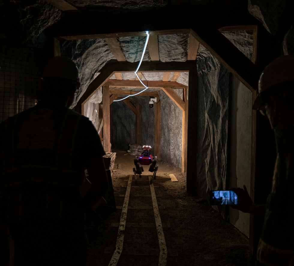 A red quadrupedal robot walks down a mine tunnel as two roboticists look on