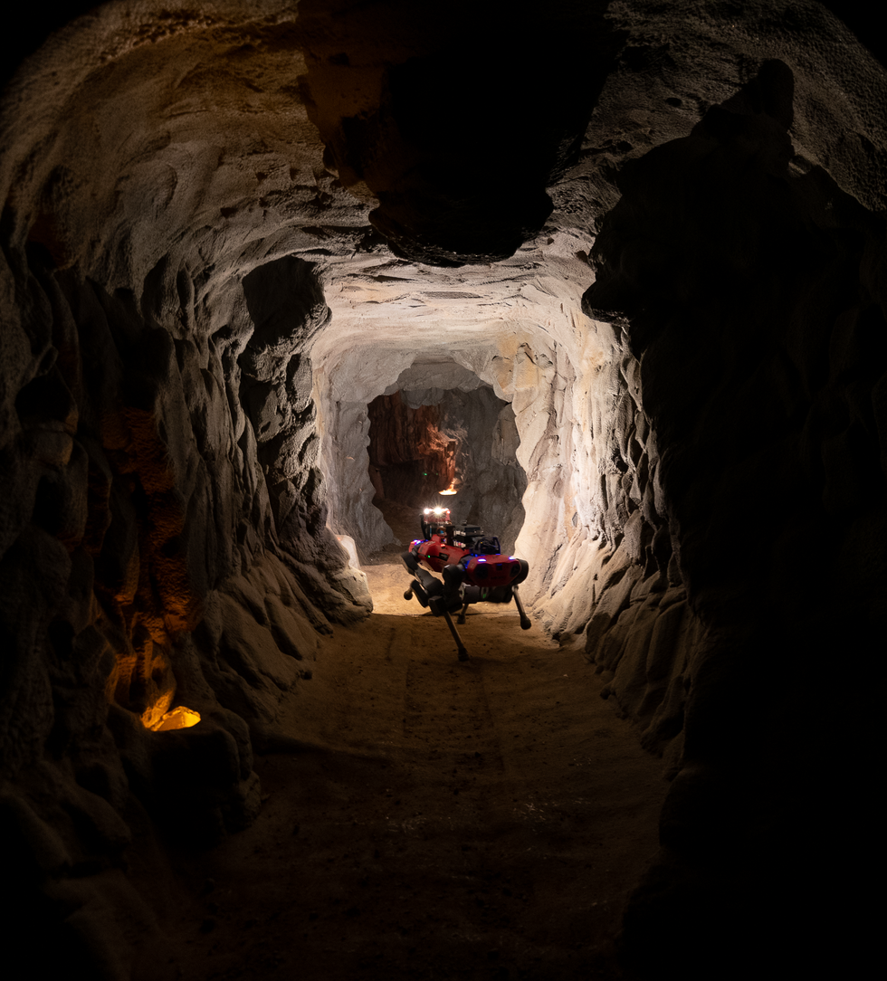 A red quadrupedal robot with a bright light explores a dark cave environment