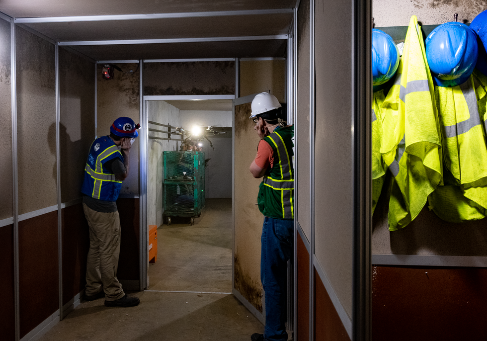 Two roboticists covering their ears watch a drone through a doorway next to a closet with yellow safety vests and blue helmets hanging on pegs