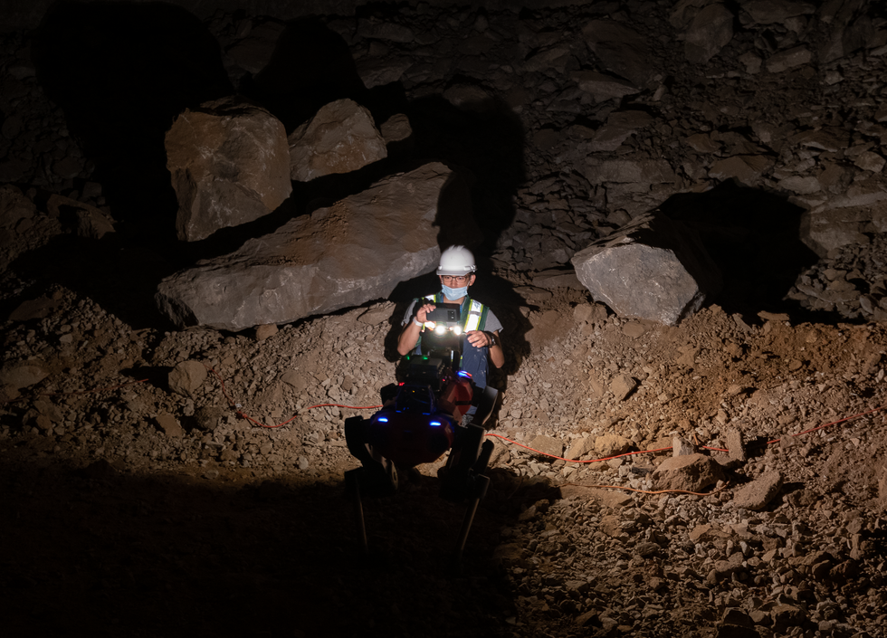 A red quadrupedal robot stands directly in front of a crouching human recording it with a cell phone in a dark cave