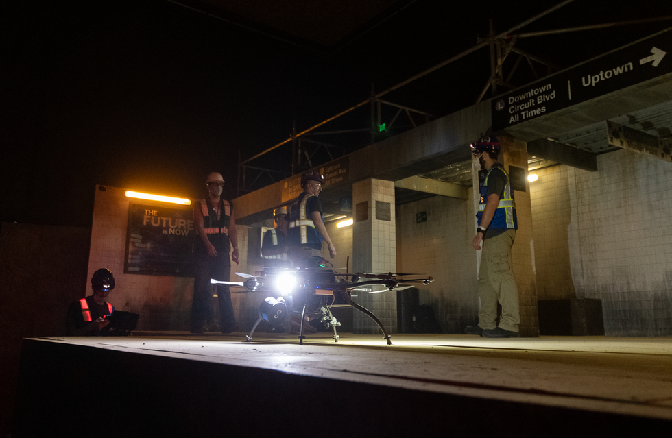 A drone with a bright light sits on a dark subway platform with a group of roboticists behind