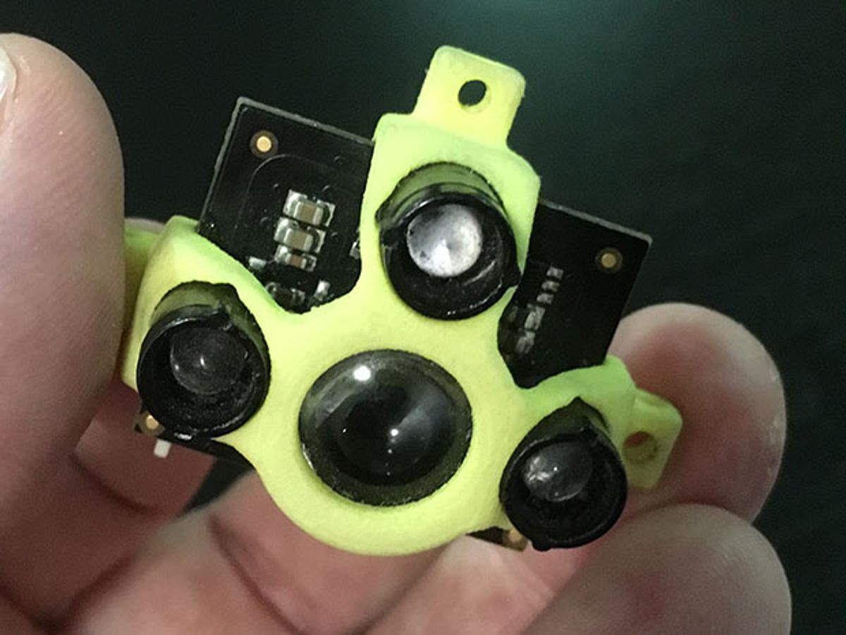 Fingers hold a small circuit board which supports four cylinders with lenses. A yellow/green piece of plastic partially covers the circuit board.