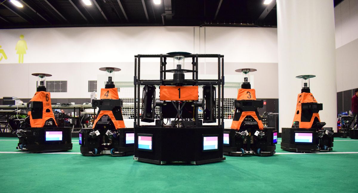 Five large orange robots with wheels line up on an indoor soccer field
