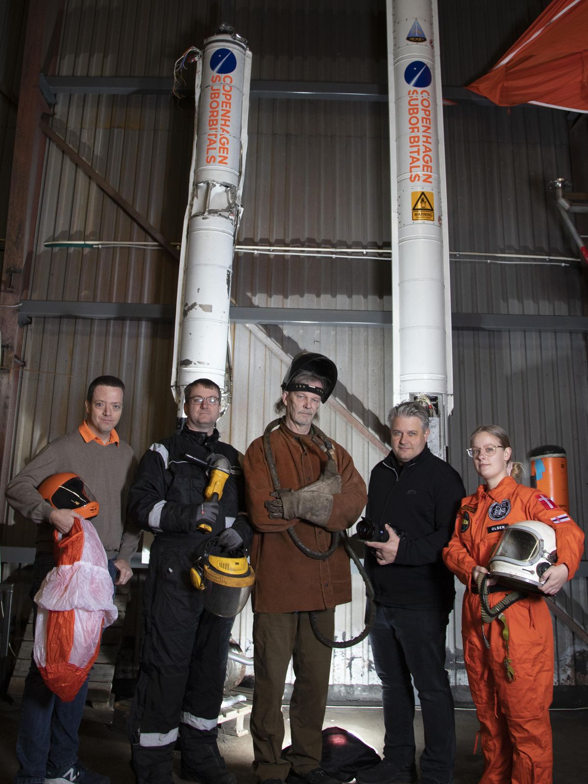 Five people stand in front of two tall rockets. Some of the people are wearing space suits and holding helmets, others are holding welding equipment.