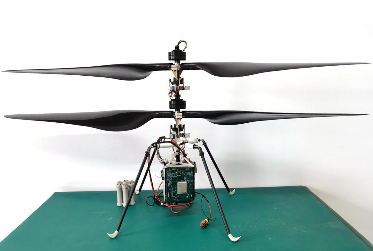 Four thin raised black legs join in an x. A vertical piece with equipment and wires is on top, and hanging under is a circuit board. The top of the base has two wide black propellers extending out.