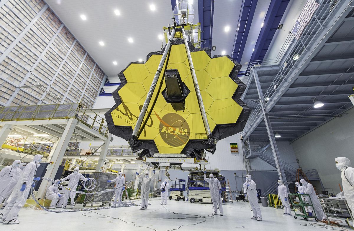 Fourteen technicians in clean-room suits guide the hoisting of a honeycombed, hexagon-mirrored telescope inside a giant cleanroom construction space 