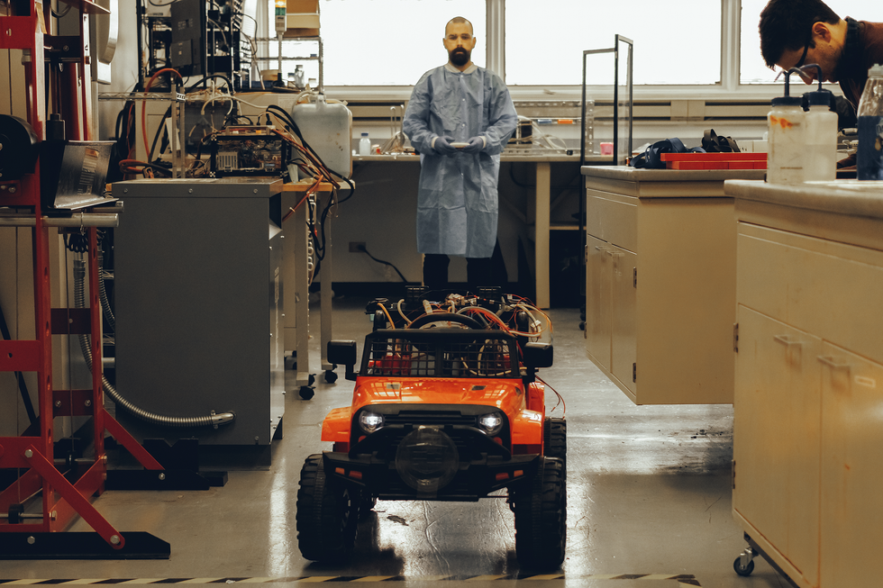 Front view of a toy jeep with black grill, orange body, open top and a mass of wires in the back. Behind the jeep stands a man in a lab coat. Various lab equipment brackets the view, and on the right side there is another man bent over a laboratory bench.