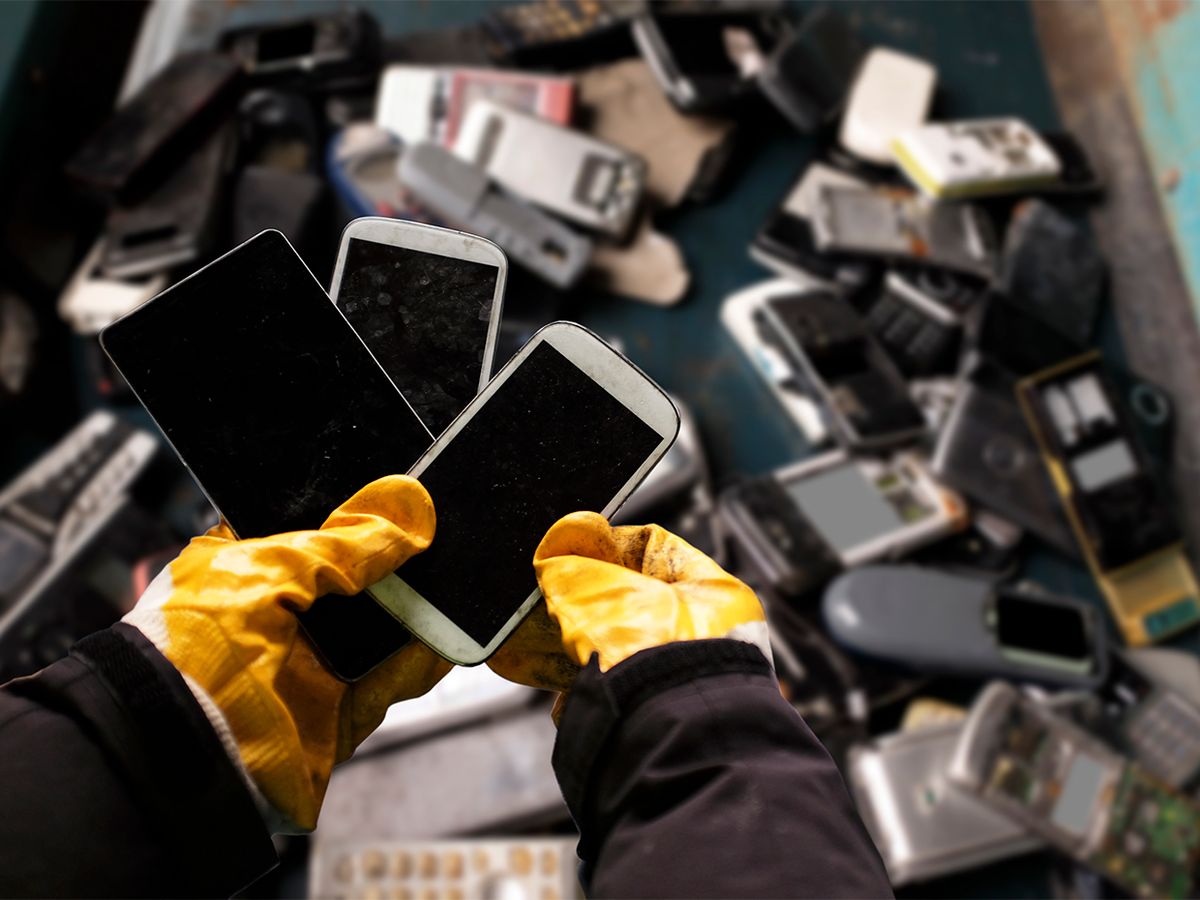 Gloved hands holding several used phones with a bin of other used electronics in the background