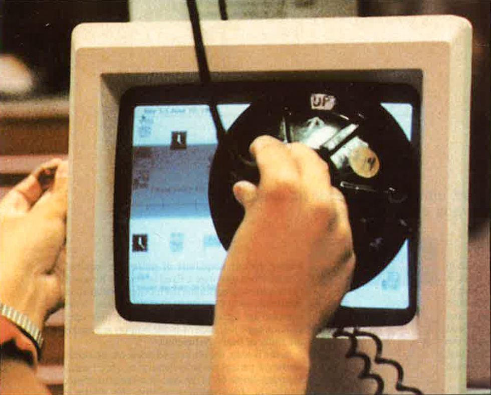 Hands hold a round cone-like object up to the front of a Macintosh computer display