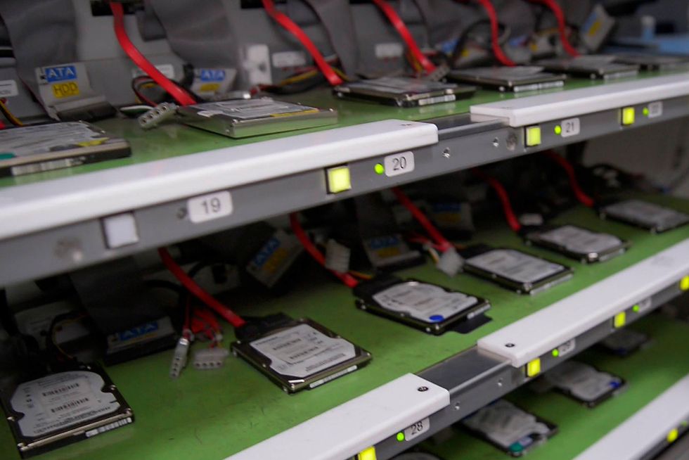 Harddrives with a red cable coming out of each sit in a row on numbered shelves