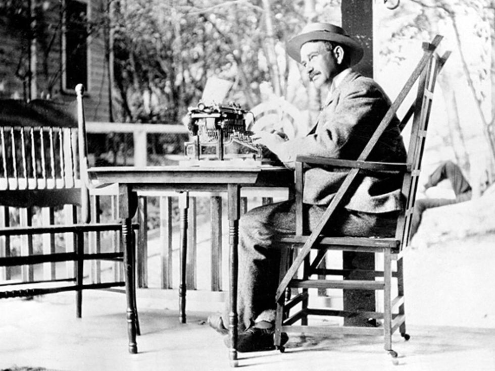  Historical black-and-white image of a mustachioed man in a hat sitting at a typewriter at a table outside.