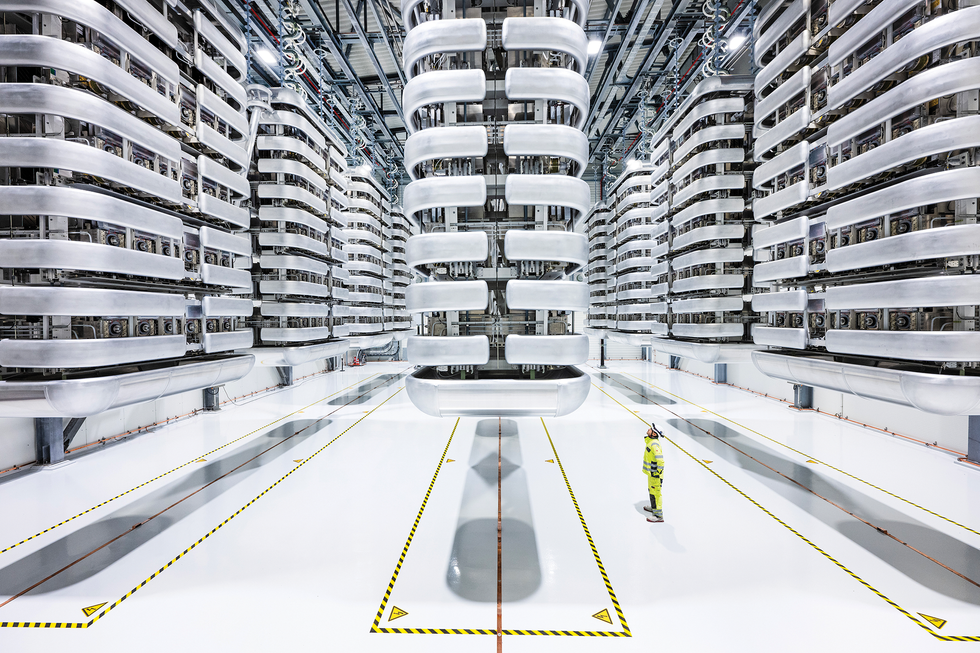 Huge, high-voltage assemblies in a brightly lit hall dwarf a worker standing amid them. 