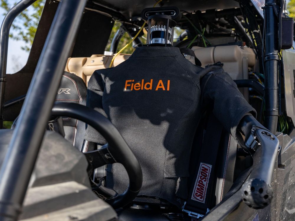 Human-like robot driver with a logo on its chest saying "Field AI" sits behind the wheel of an open, dune buggy-like vehicle