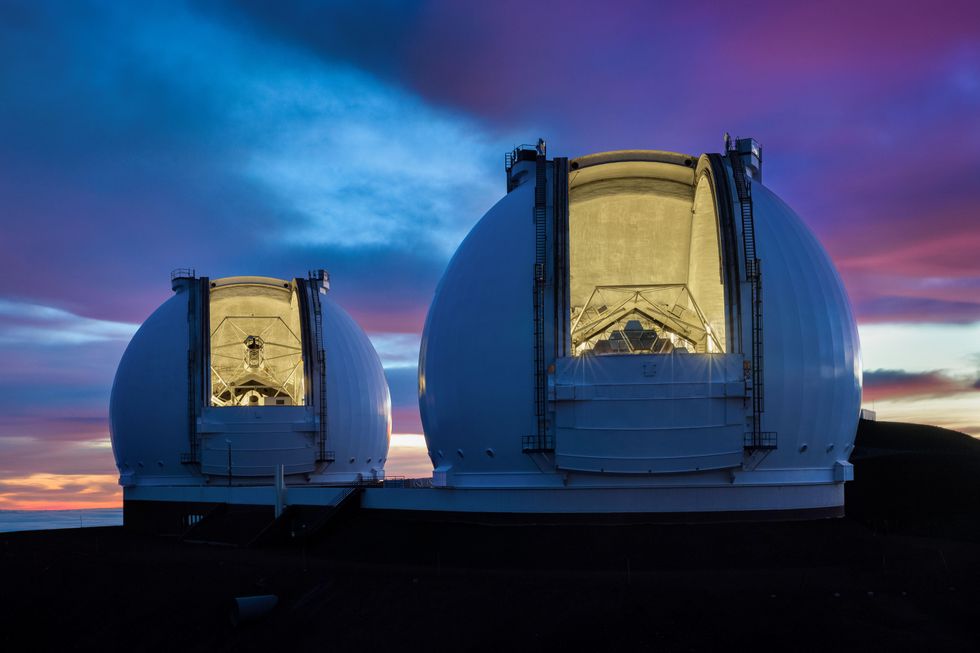 Image of two giant telescope domes opened to reveal big telescopes inside, the Keck I and Keck II telescopes; outside is a cloudy night at sunset