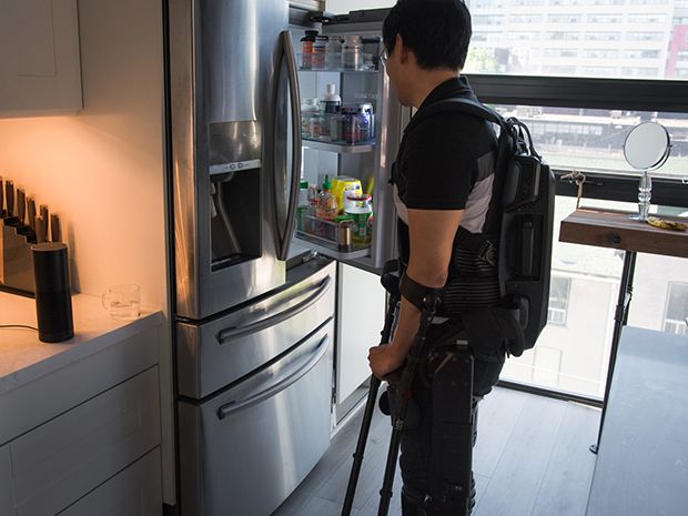A man wearing a robotic exoskeleton stands in front of the fridge; the Amazon Echo sits on the counter nearby.