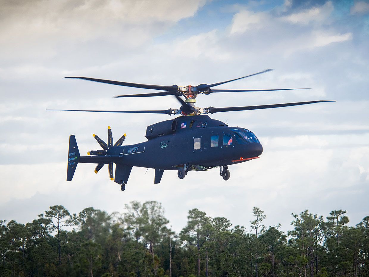 The black Defiant helicopter hovers above the treetops against a cloudy blue sky in a photo taken during its maiden flight.