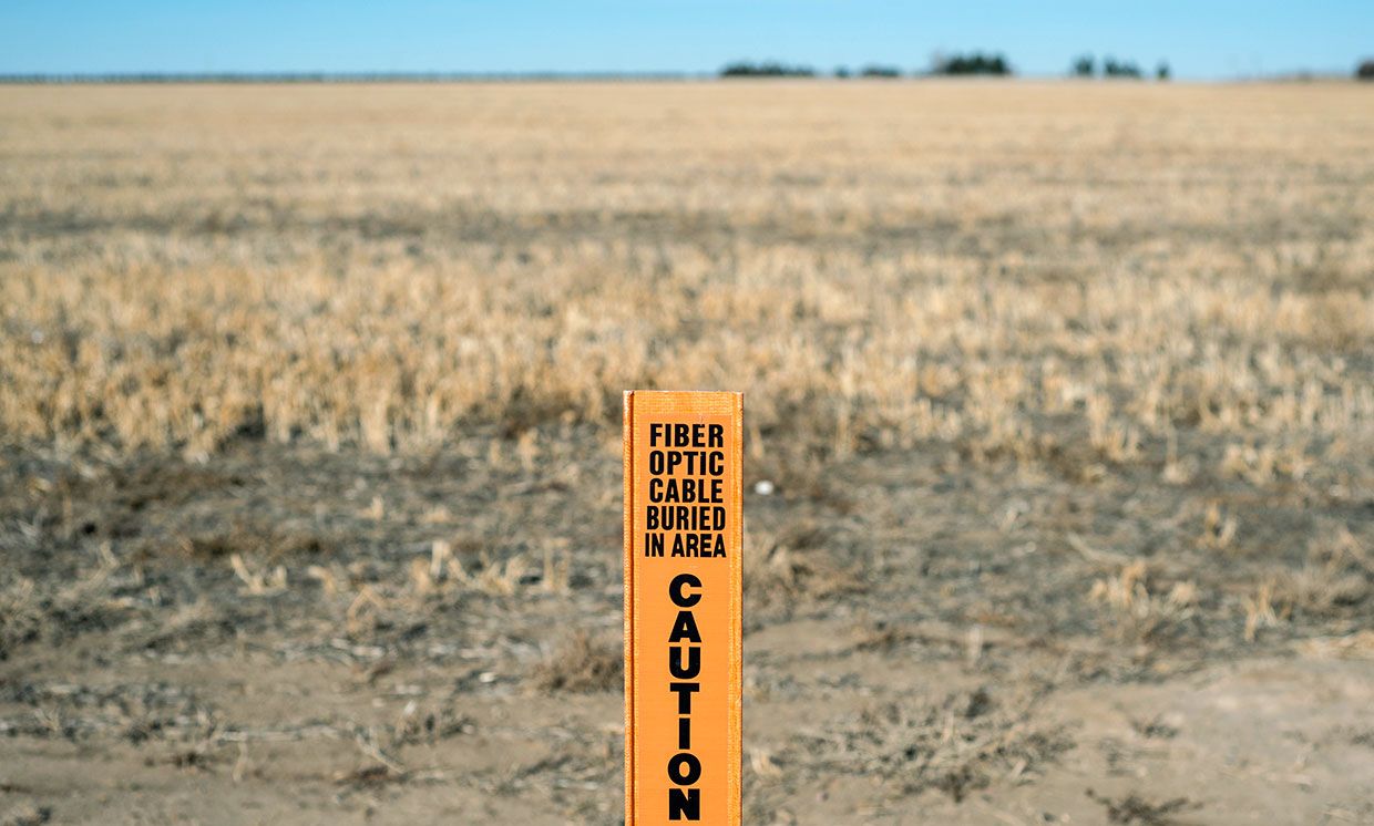 Photograph of a sign saying "Fiber optic cable buried in area, caution" in the foreground, with farmland in the background.