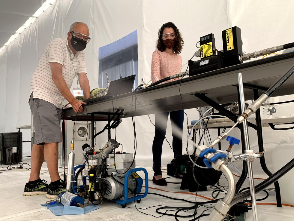 A man and a woman wearing masks stand at a table in a white tent. In the foreground is silver and blue equipment including a nozzle from which white spray is emitting.