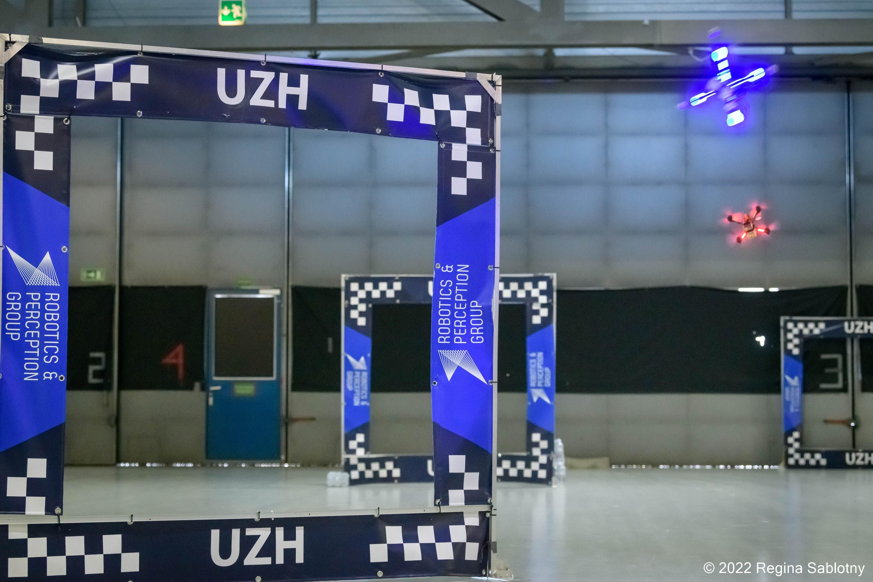Two drones, one red and one blue, fly through gates in a drone racing course inside an aircraft hangar
