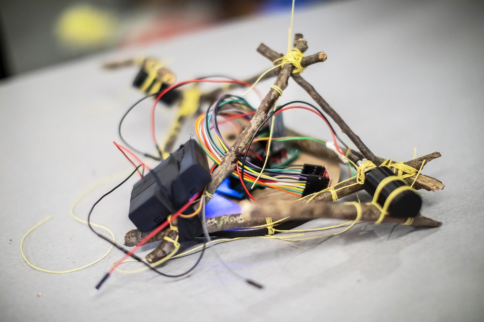 An image of a robot made of a small sticks tied together with a tangle of colorful wires, batteries, actuators, and electronics
