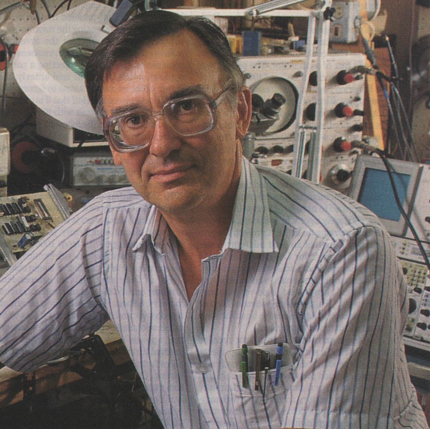 Man in white striped shirt with pocket protector seated in front of oscilloscope and other electronics equipment