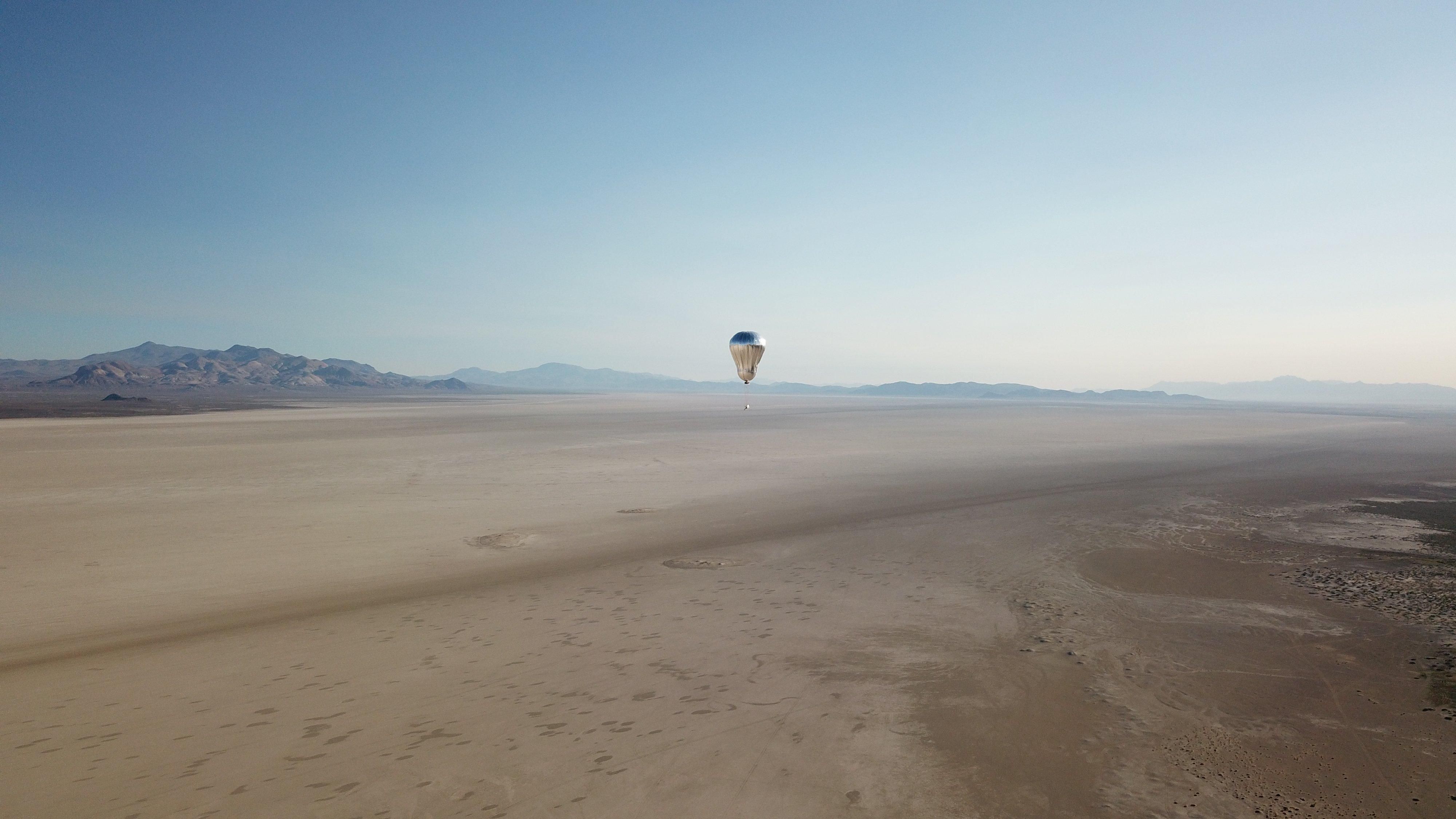 A silver balloon with a robotic payload drifts high above a desert landscape
