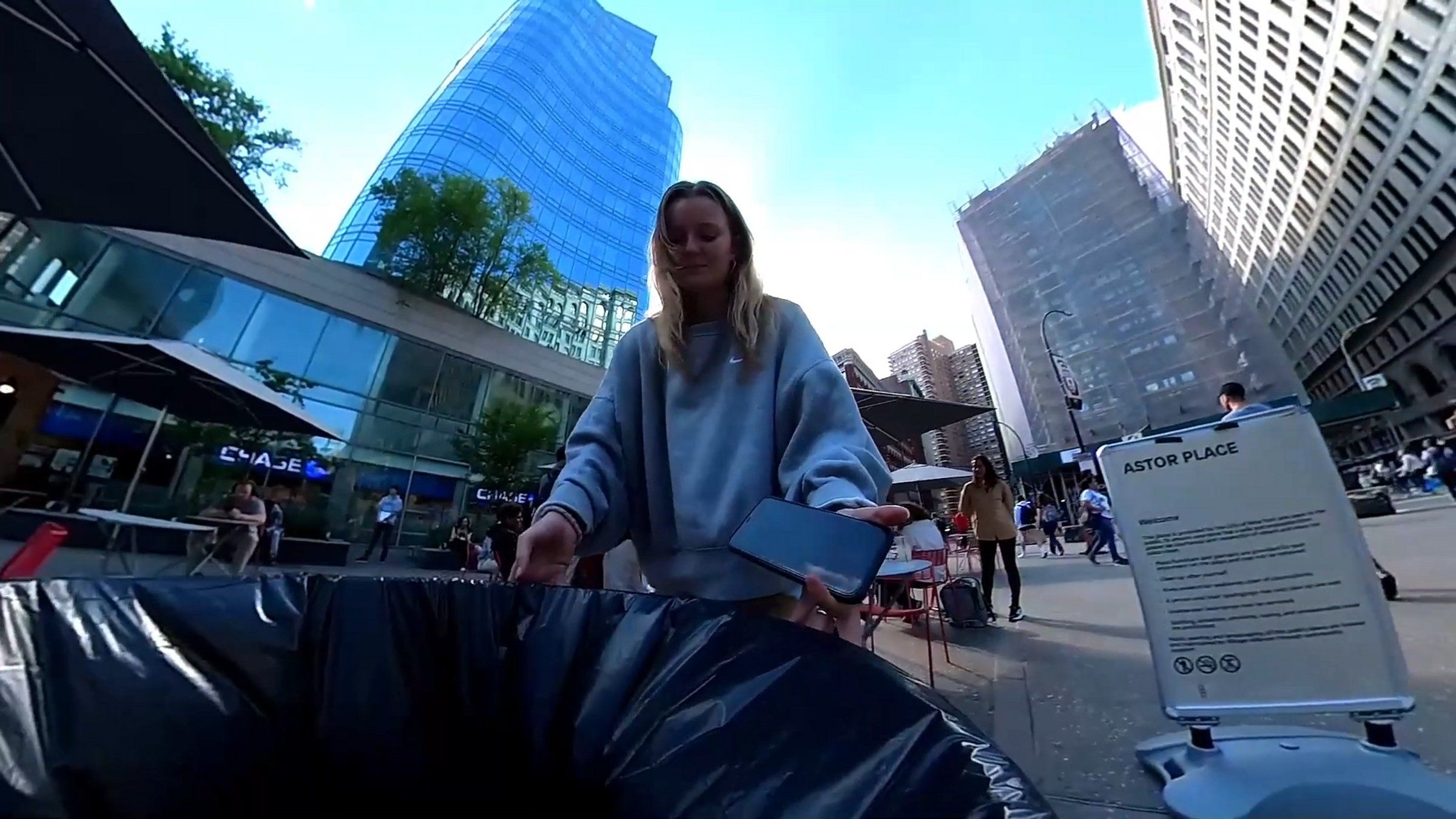 A video screen capture showing a NYC street and a woman helping a trash barrel robot over a bump
