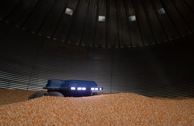 A small blue robot with linear corkscrew wheels sits on a pile of grain in a huge dark grain bin