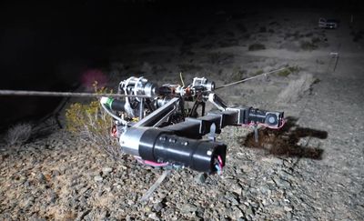 A spindly robot made of cables and actuators slowly travels down a cable above a desert landscape.