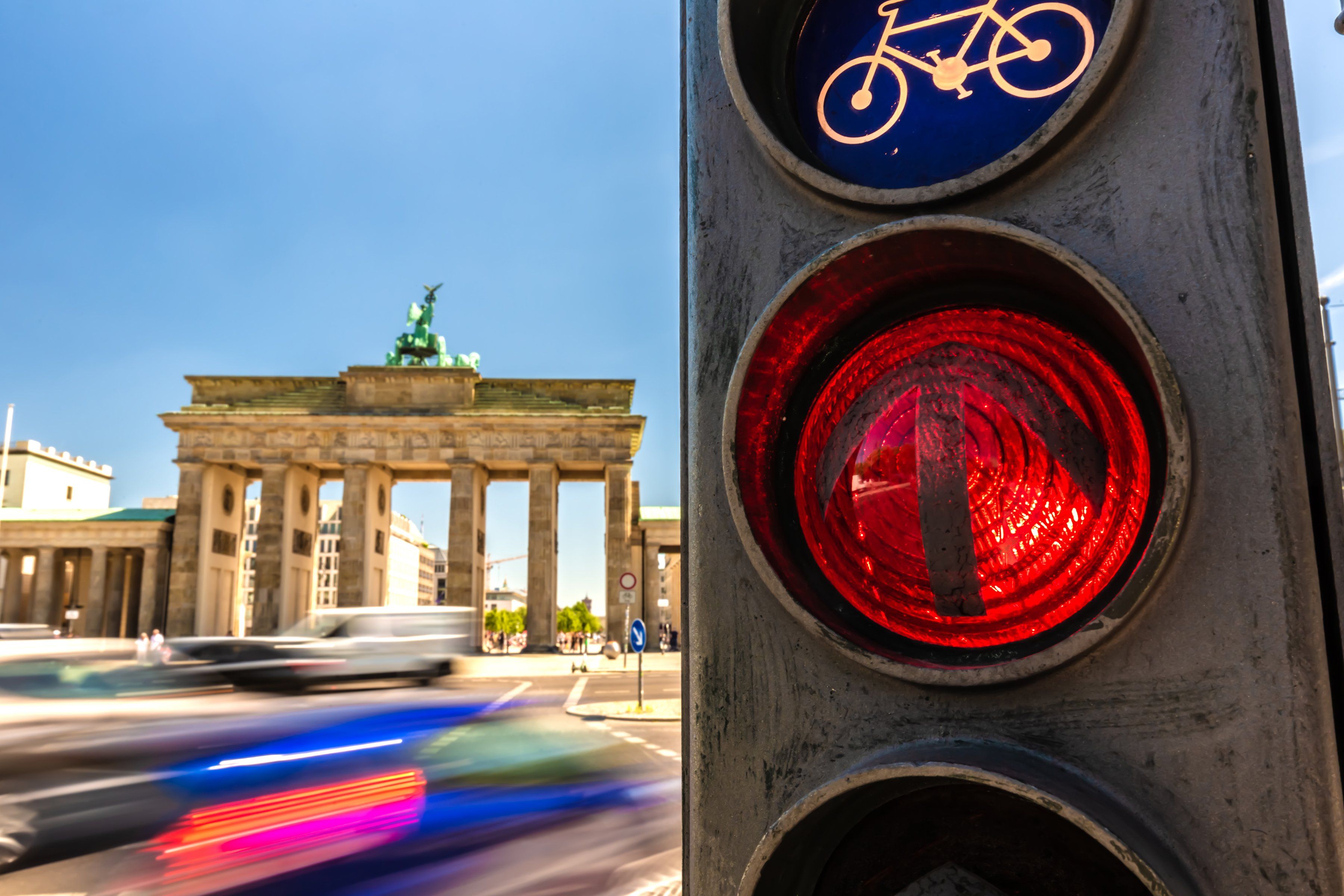 In the foreground, a German traffic light is lit red for cars and go for bikes. In the background, motion blurred traffic moves through a busy intersection.