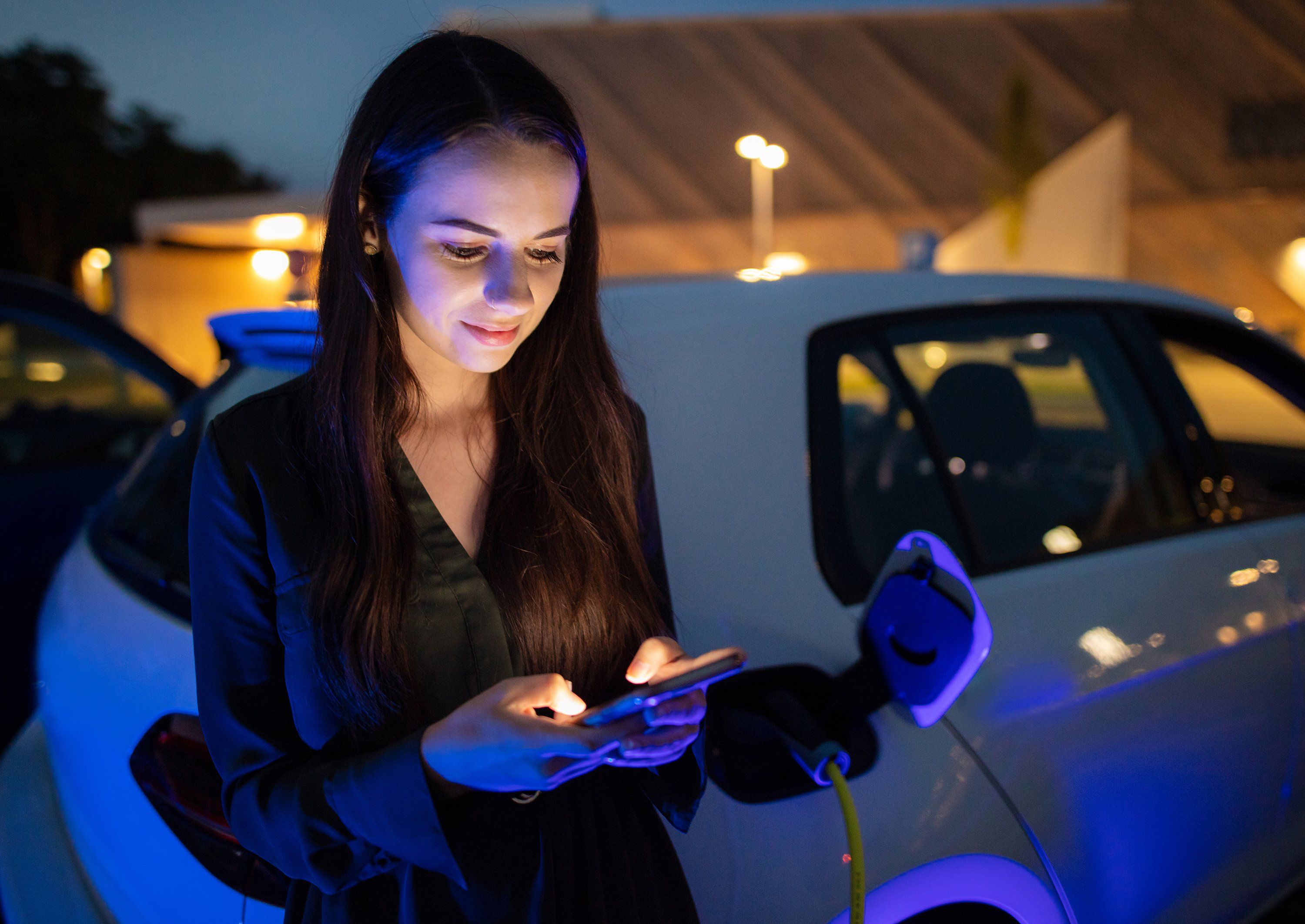 Nighttime photo of a woman looking at her phone as her electric vehicle charges.