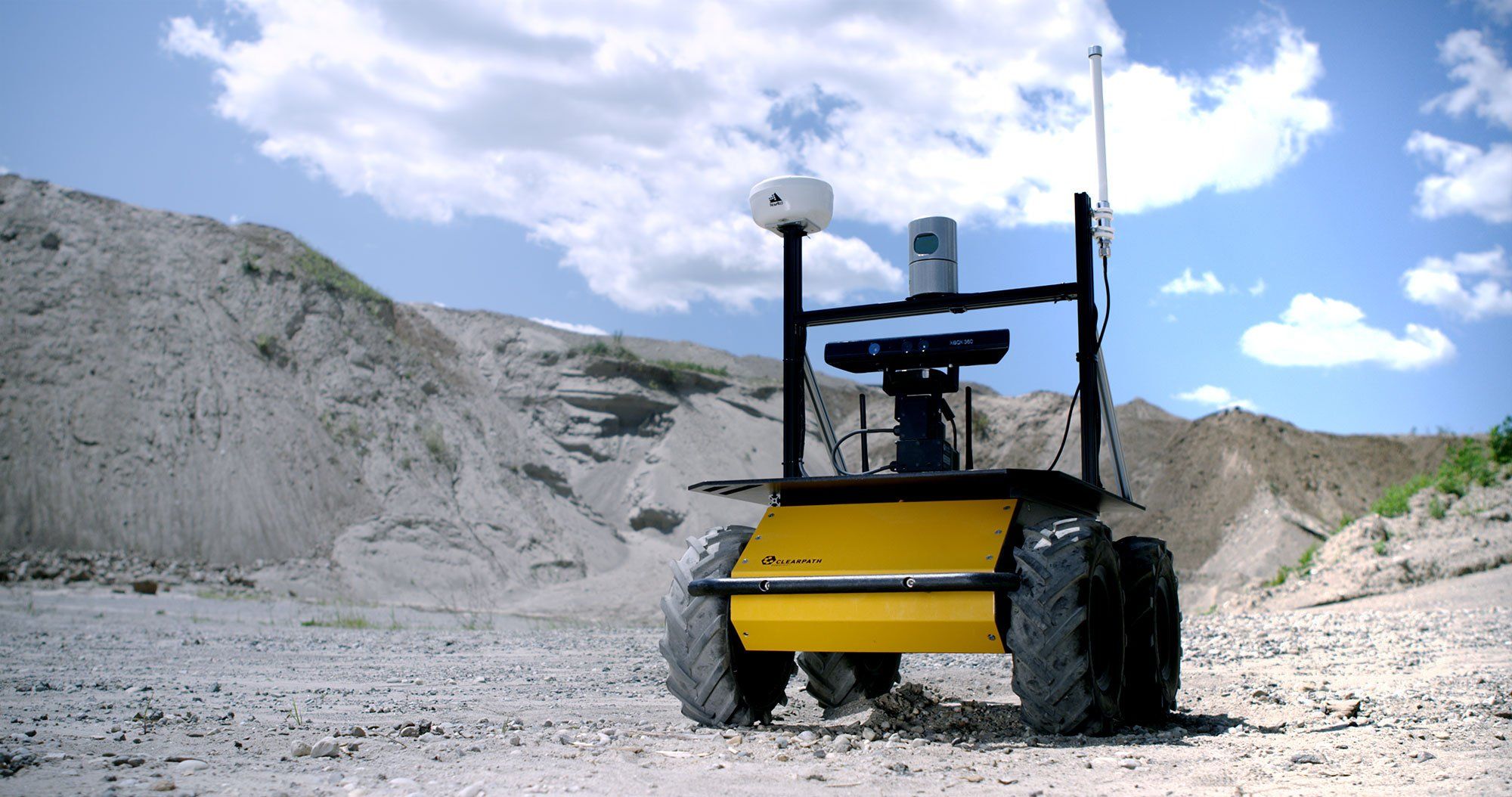A photo of a small yellow body robot vehicle with black wheels and sensors on top patrolling a quarry.