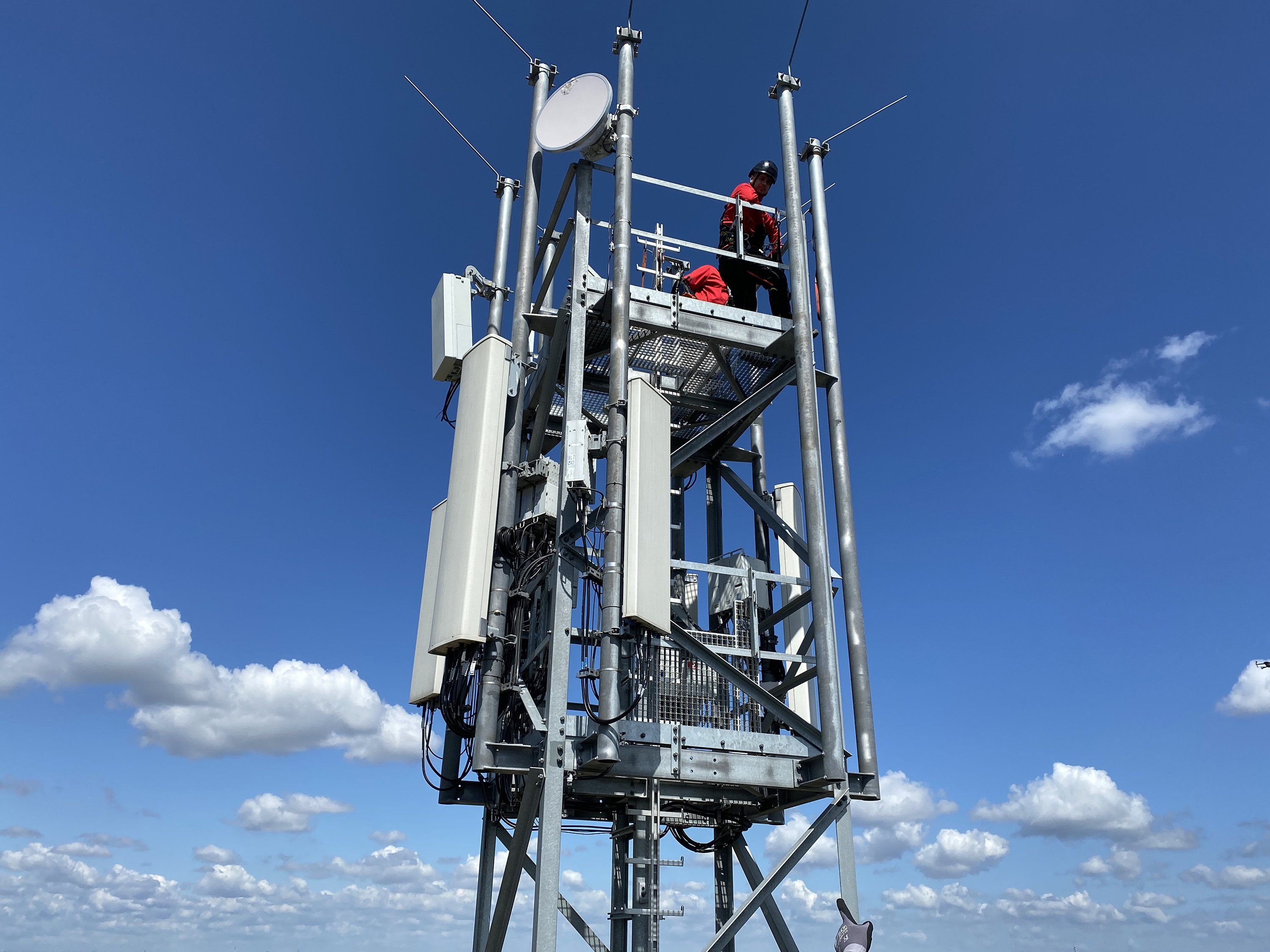 Two helmeted people stand at the top of a communications tower with 5G massive MIMO multi-antenna systems. A bright blue sky with clouds is behind them.