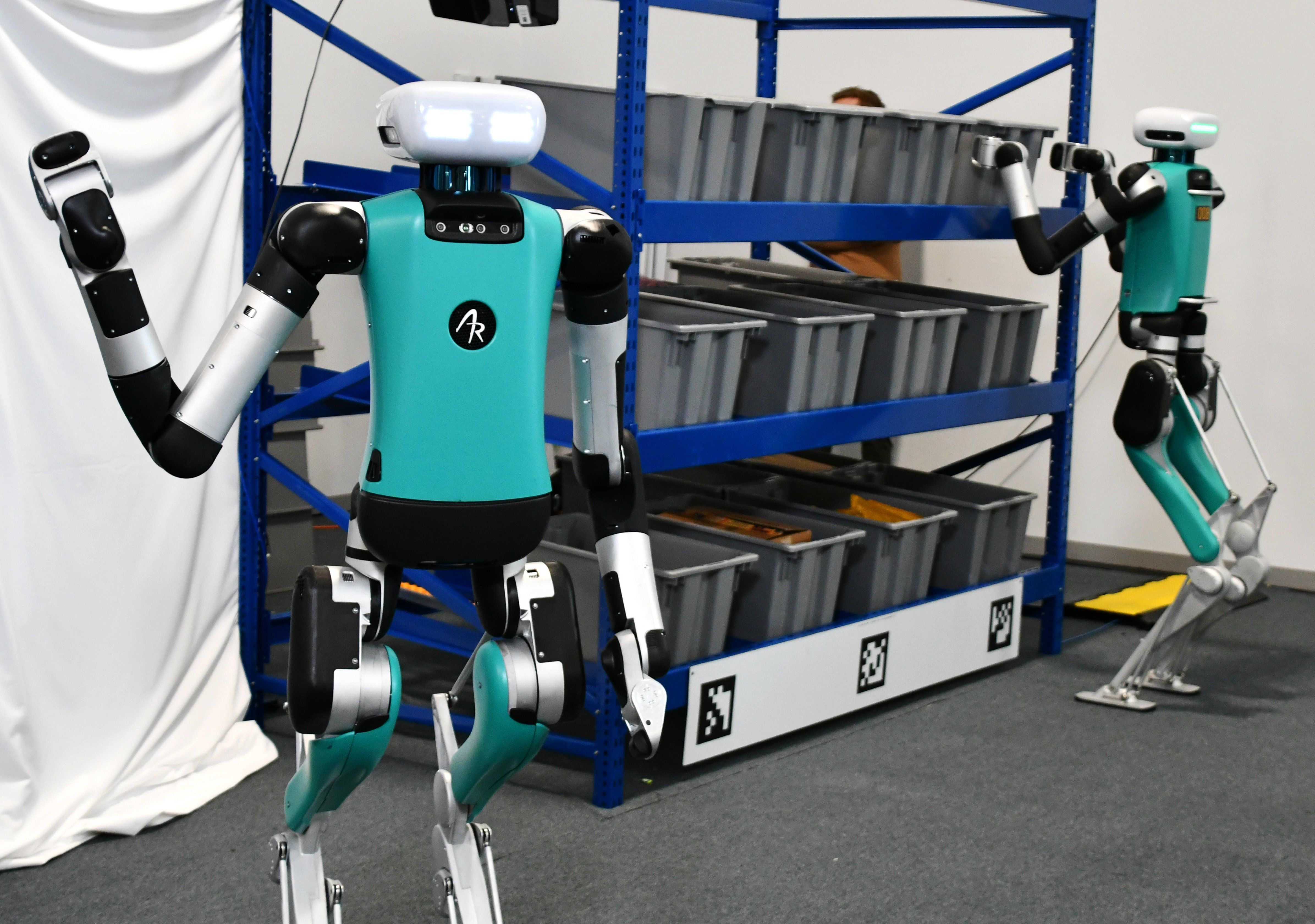 In the foreground, a teal, black, and silver bipedal humanoid robot waves its right arm. In the background, a similar robot reaches to take a bin off of a shelf.