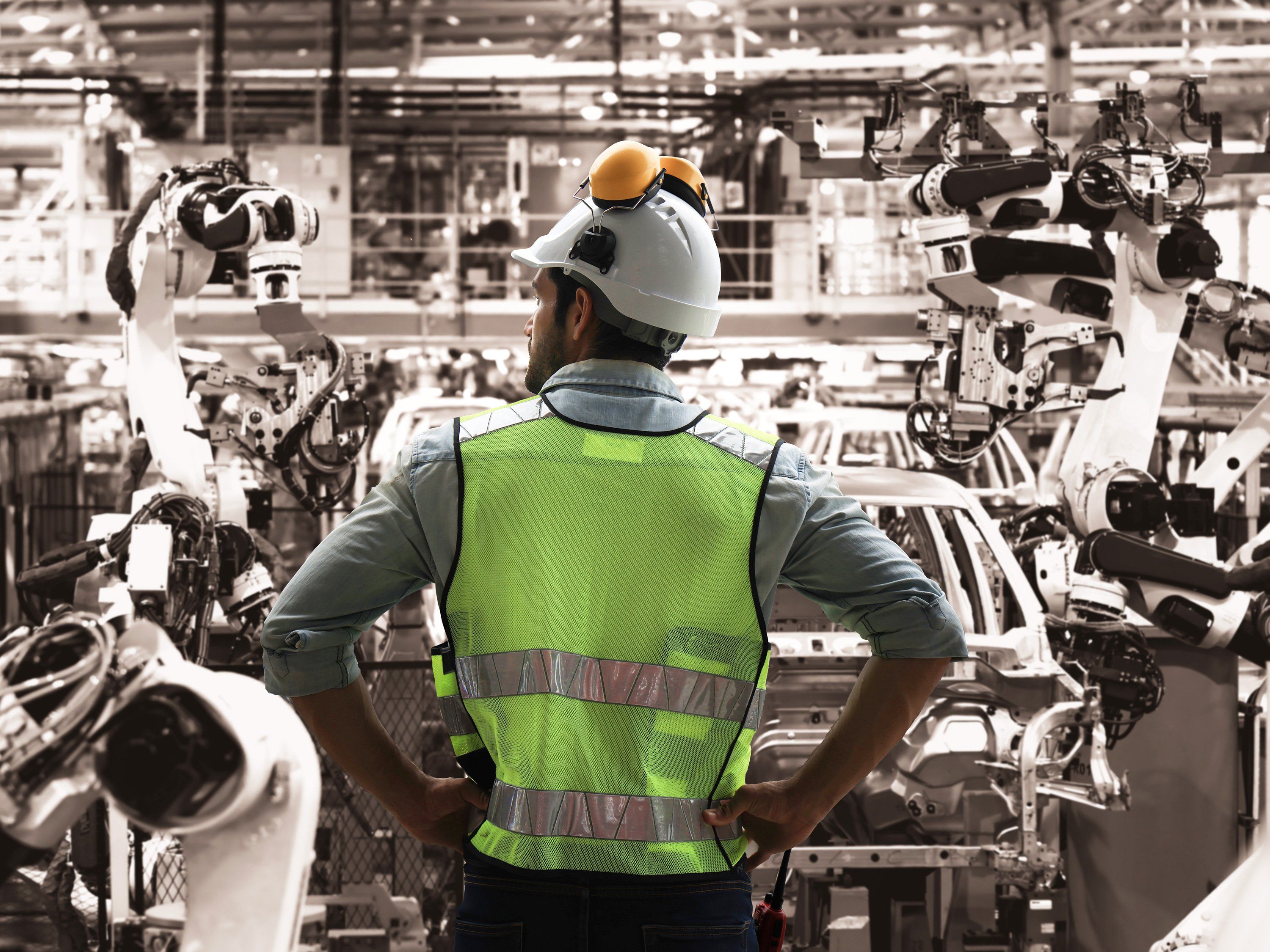 man standing with back to camera with yellow vest and hard hat looking out over a manufacturing plant