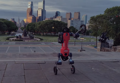 A red bipedal robot with wheels for feet and hands stands upright at the top of steps with the city of Philadelphia in the background