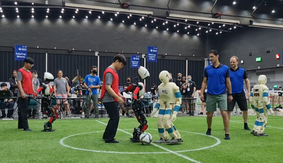 Humans and human-size humanoid robots stand together on an indoor soccer field at the beginning of a game
